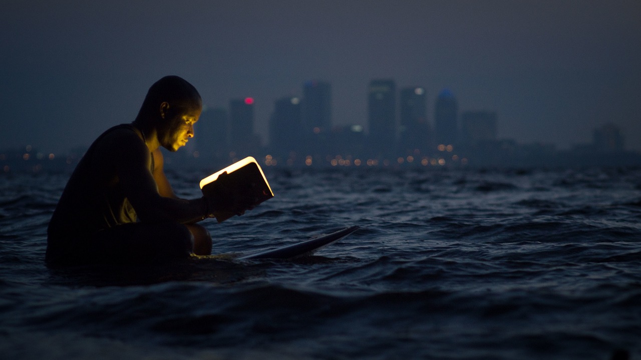 man sitting on surfboard at sea reading an ebook